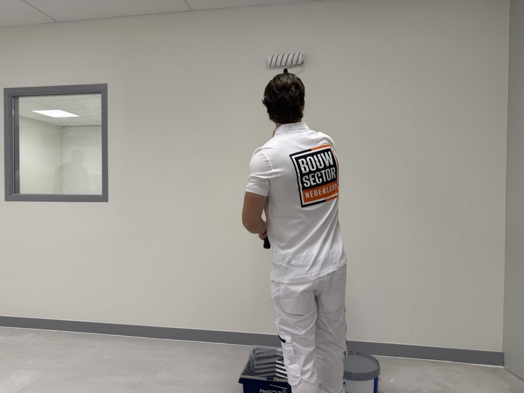 Schilder in witte werkkleding rolt een lichte wand naast een binnenraam, met verfbak en emmer aan zijn voeten