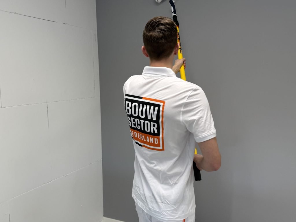 Schilder met het logo van Bouwsector Nederland op zijn rug zet een roller op een lange steel tegen een grijze wand