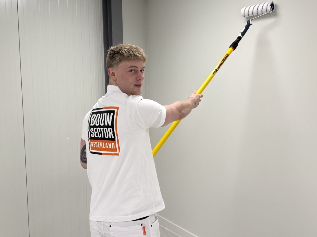 Schilder kijkt over zijn schouder terwijl hij met een verlengsteel een witte muur rolt