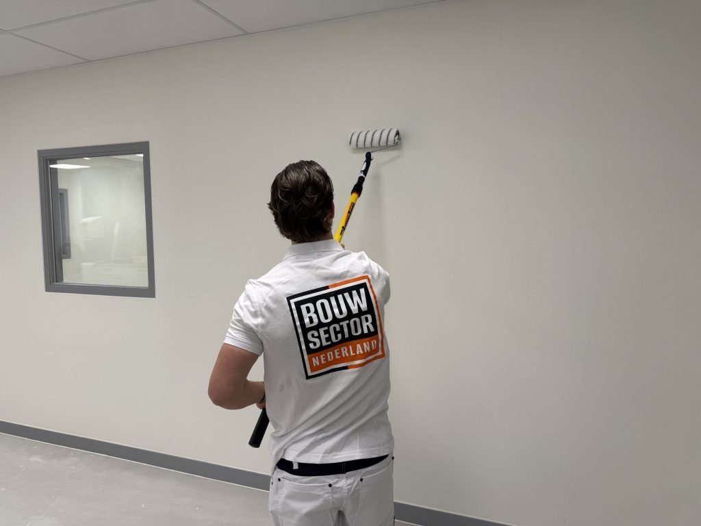 Schilder met het Bouwsector-logo rolt een lichte wand in een lege ruimte met een binnenraam en een grijze plint
